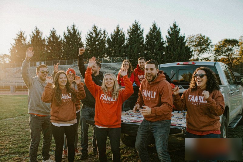 A group of friends tailgating at a football game by a pickup truck bed, wearing matching personalized team hoodies, with a custom picnic blanket spread out